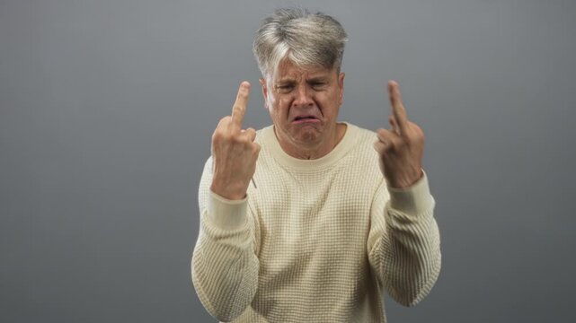 Man with grey hair wearing cream sweater holding car keys and showing middle fingers against a studio gray backdrop, pained grimace; frustration.