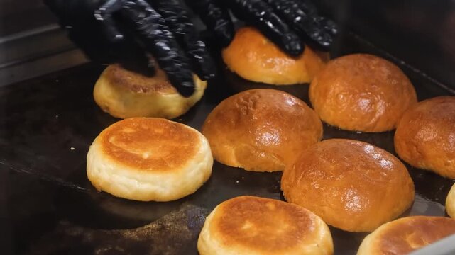 Close-up footage of several golden-brown burger buns toasting on a professional black griddle or baking tray in a commercial kitchen or bakery setting.