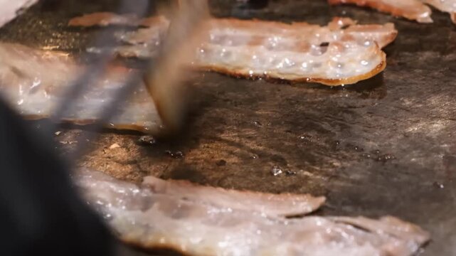 A close-up shot capturing several strips of pork bacon as they fry and sizzle in their own fat on a hot flat-top griddle. The footage highlights the Maillard reaction as the meat browns and the fat re