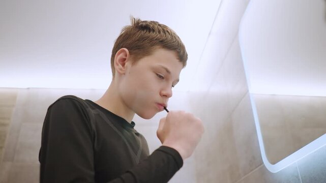 Youth grooming routine. Young boy preparing for day ahead. Child diligently brushing teeth in small bathroom mirror. Teenager concentrating on dental hygiene before heading out for sports activity