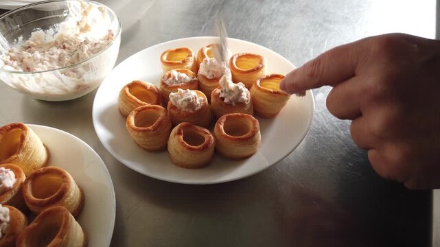 A hand arranges golden puff pastry shells filled with a creamy mixture on white plates beside a glass bowl in a stainless steel kitchen, evoking casual entertaining, hors d'oeuvres preparation and pla