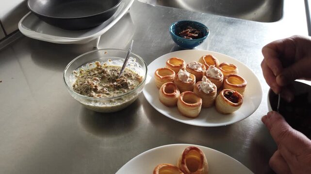 A hand arranges golden puff pastry shells filled with a creamy mixture on white plates beside a glass bowl in a stainless steel kitchen, evoking casual entertaining, hors d'oeuvres preparation and pla
