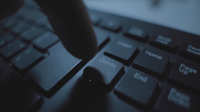Close up on computer keyboard as hand presses delete keypad in dark room. Office or home. Secret agent desk erasing data or files from computer or server. Man clicks on button to remove file to trash