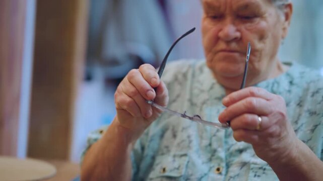 Elderly caucasian grandma picks up glasses from table placemat, lifts frames to face with concentrated expression, warm kitchen light, teapot and biscuits nearby, small domestic victory moment.