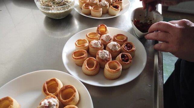 A hand arranges golden puff pastry shells filled with a creamy mixture on white plates beside a glass bowl in a stainless steel kitchen, evoking casual entertaining, hors d'oeuvres preparation and pla