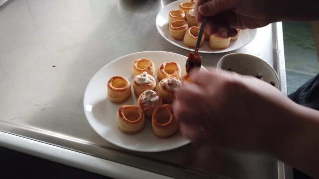 A hand arranges golden puff pastry shells filled with a creamy mixture on white plates beside a glass bowl in a stainless steel kitchen, evoking casual entertaining, hors d'oeuvres preparation and pla