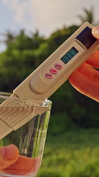 First Person View Of Hands Measuring Purity Of Drinking Water With Digital Tester Showing Twenty Ppm In Clear Glass Against Tropical Green Landscape. Clean Drinking Water Verification
