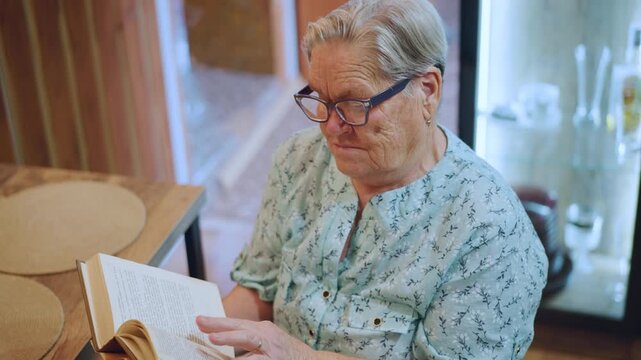 Grandmother enjoying her book. Senior lady peruses her favorite novel peacefully. Warmly dressed grandmother attentively reads cherished book at her kitchen table surrounded by comforting items
