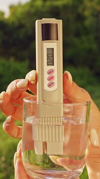 Close View Of Female Hand Holding Glass With Digital Water Quality Tester Showing Twenty Ppm Ppm Measurement Against Tropical Green Landscape. Drinking Water Safety Monitoring