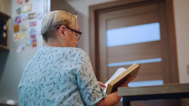 White grandma reading memoir at counter, elderly white woman with glasses sits at kitchen counter holding open book, fridge with magnets and wooden door in background, warm afternoon light, retiree