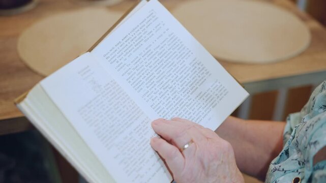 Elderly lady studies open pages quietly. White elder woman siting at table immersed in reading activity. An aged white grandmother peacefully reads book at her dining table during daylight hour