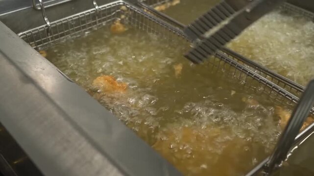 Close-up footage of marinated chicken pieces being deep-fried in hot oil inside a commercial kitchen deep fryer. The chicken sizzles as it cooks to a crispy golden brown, perfect for food preparation,