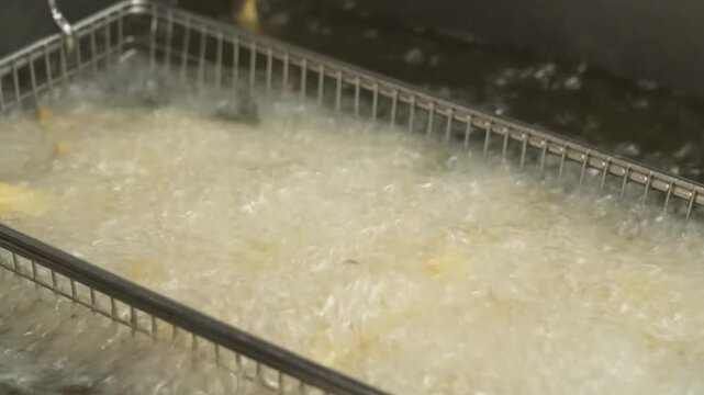 close-up shot captures golden French fries sizzling and bubbling in hot oil inside a deep fryer basket. The video highlights the texture and cooking process of the crispy fast food snack, perfect for 