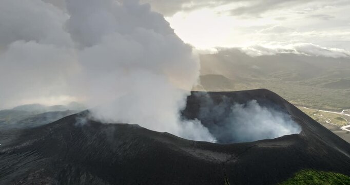 Aerial panorama of Mount Yasur volcano in Vanuatu's Tanna Island actively erupting, smoke plumes rising from its dark crater against a cloudy sky with surrounding green landscape and a winding river