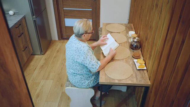 Elderly woman sorting mail at kitchen table, opening envelopes and checking bills, focused expression, mug and jar nearby, tidy domestic counter, practical routine and quiet concentration