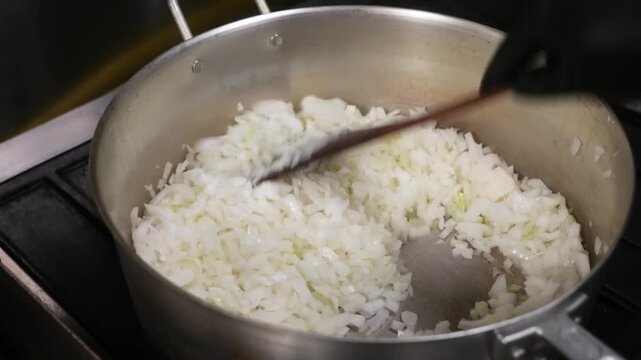 High-angle, close-up shot of finely diced white onions being stirred and saut&eacute;ed in a large stainless steel stockpot. The onions are becoming translucent, showing the initial stages of a cooking proce