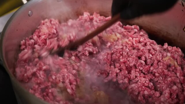 Close-up shot of a hand in a black glove using a wooden spoon to stir raw ground beef in a stainless steel pot or pan. The meat is in the process of browning and cooking, likely for a dish such as bol