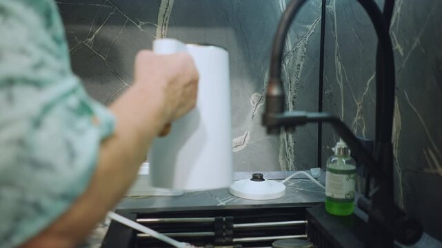White grandmother hands placing white kettle at sink for drying, steady faucet and marble backsplash visible, careful wipe and reposition, calm domestic cleaning routine, retired homemaker tending