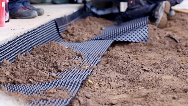 Worker laying down drainage membrane on soil surface, with dirt piles and construction tools visible in the background, demonstrating a methodical installation process