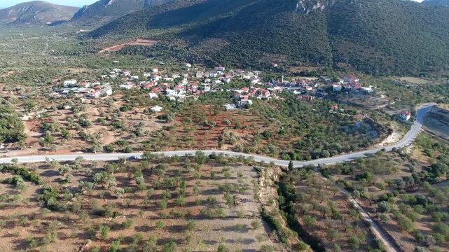 Aerial view captures hilltop village in Italy surrounded by fruit orchards on Mediterranean slopes. Panoramic scene reveals rural hamlet amid citrus groves, coastal hillsides, farmland landscape.
