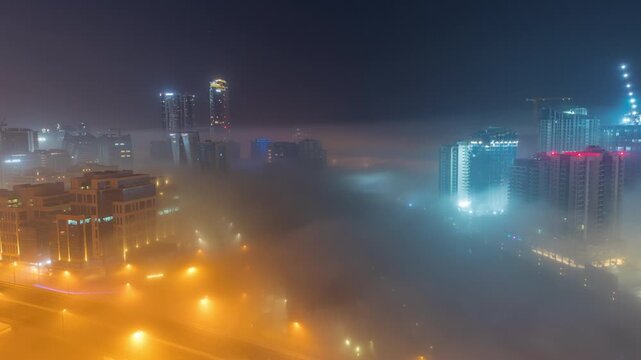 Dubai Business Bay canal at night timelapse, aerial top view. Illuminated skyscrapers and cranes covered in thick glowing fog layer over water canal, colorful urban nightscape, UAE.