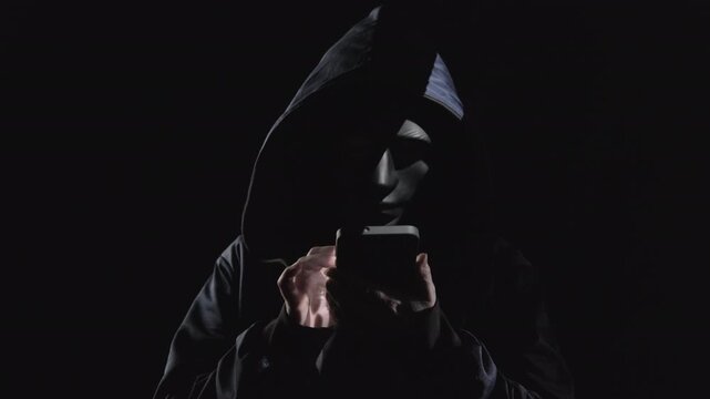 Man hiding face behind black mask dials number on smartphone and makes call, phone scam, voice phishing