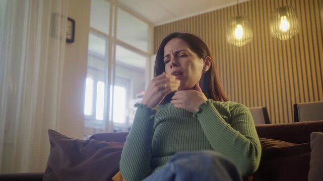 Symptoms from flu season in winter concept. Close-up of unhealthy young caucasian woman sitting on couch at home touching her neck and suffering from sore throat, angina, hard to swallow, voice loss 