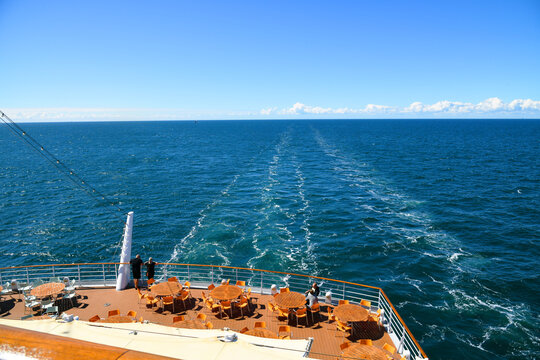 The view from the stern deck of a cruise ship to the open sea and the lane of the ship.
 The rear deck is equipped with numerous tables and chairs for passengers.