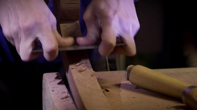Woodworker using a metal scraper to refine the shape of a curved timber piece on a workbench. Slow motion close up.