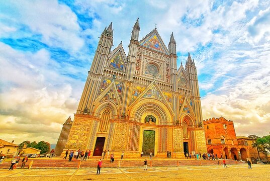 Magnificent Gothic Facade of Orvieto Cathedral (Duomo di Orvieto) with golden Mosaics and tourists under cloudy blue sky.