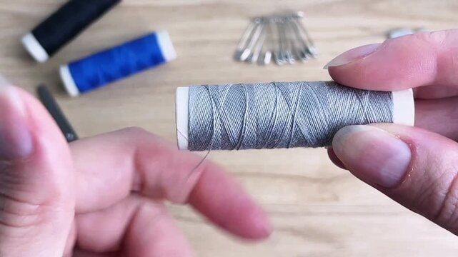 A close-up of hands unwinding gray thread from a spool on a workbench with sewing tools. Conceptual reference to handicrafts, thread preparation for sewing, and clothing repair.