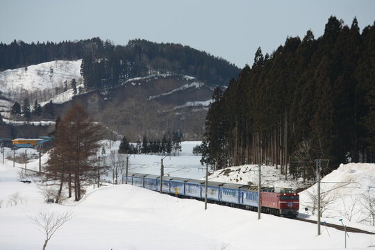 雪の積もる冬の山間部を走行する赤い電気機関車と寝台列車