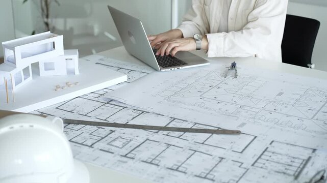 Architect working on new residential project designs, using laptop and reviewing blueprints with an architectural model and safety helmet on planning table
