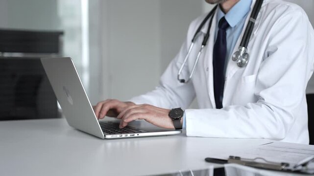 Confident doctor wearing a white coat and stethoscope, using a laptop at a desk for medical record keeping and online patient care. Medicine, healthcare and science concept