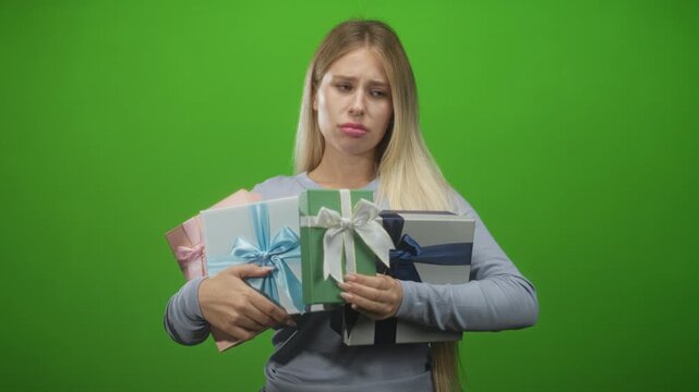 Young blonde woman holding multiple wrapped gift boxes with hands clasping ribbons and pouting lips in studio green screen, wearing long sleeve top; disappointment.