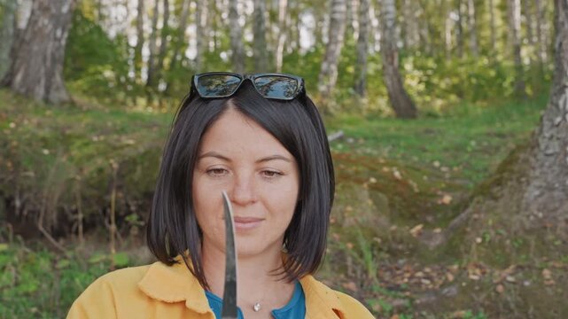 Caucasian woman camping performing safety blade check near camp studies knife edge with thoughtful expression, yellow jacket, sunglasses on head, quiet birch grove, readiness mood before cooking