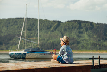 pensive young boy wearing straw hat and plaid shirt sitting on wooden pier looking at sailboat on calm lake as concept of childhood dreams, peaceful solitude and summer vacation in countryside