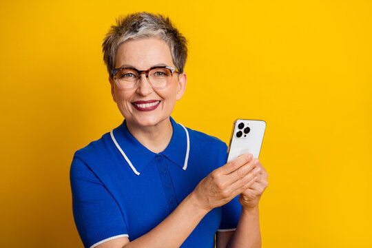 Mature woman with short gray hair wearing glasses smiles and holds a smartphone against a bright yellow background