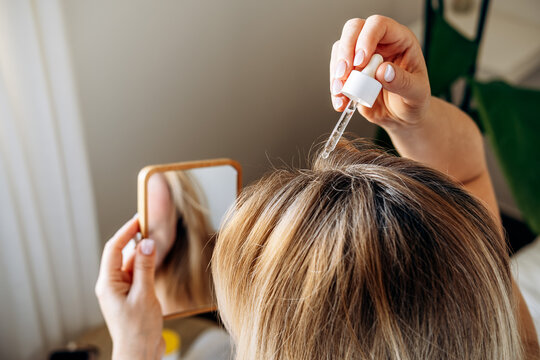 Woman applying hair serum with dropper to scalp while lying on a bed, focusing on hair care and beauty routine