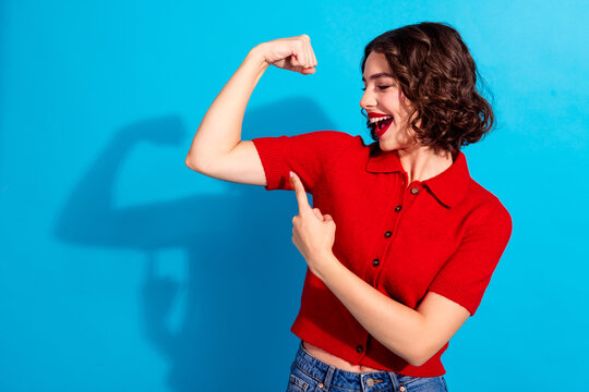 Young chic woman in red polo shirt flexing arm and smiling against blue background showcasing fashion confidence and style
