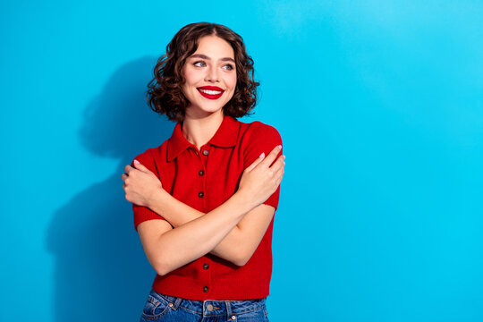 Young chic woman in red polo poses with arms crossed against blue background smiling brightly