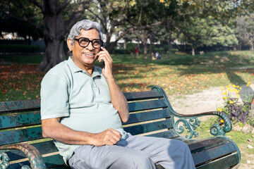 Fototapeta premium Senior Indian man talking on smartphone while sitting on a park bench. Concept of communication, connection, and modern elderly lifestyle.