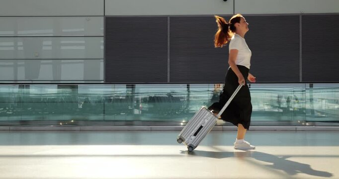 Stylish passenger woman races through modern airport terminal, effortlessly pulling her rolling suitcase behind her as she hurries to catch final call. Captured in side-view slow-motion shot