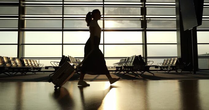 Passenger woman walk confidently with wide stride, pulling wheeled suitcase behind, silhouette shot at airport terminal. Low sun shine through window on background, camera turn right to follow woman