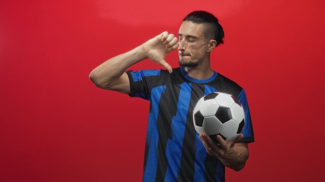 Man in blue striped jersey holding ball in left hand and showing right thumb pointing down in red studio; disapproval critique.