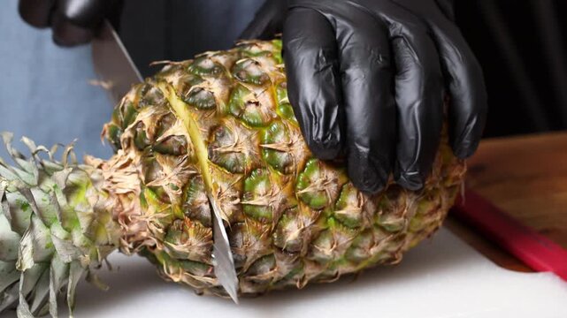 A chef wearing black gloves cuts pineapple with a knife on a cutting board.
