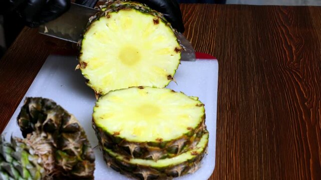 A chef wearing black gloves cuts pineapple with a knife on a cutting board.