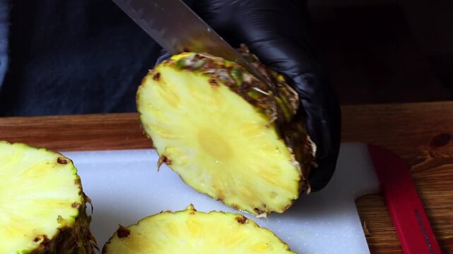 A chef wearing black gloves cuts pineapple with a knife on a cutting board.