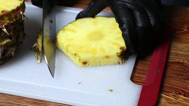 A chef wearing black gloves peels a pineapple with a knife on a cutting board.