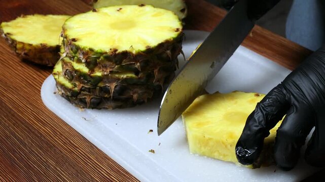 A chef wearing black gloves peels a pineapple with a knife on a cutting board.